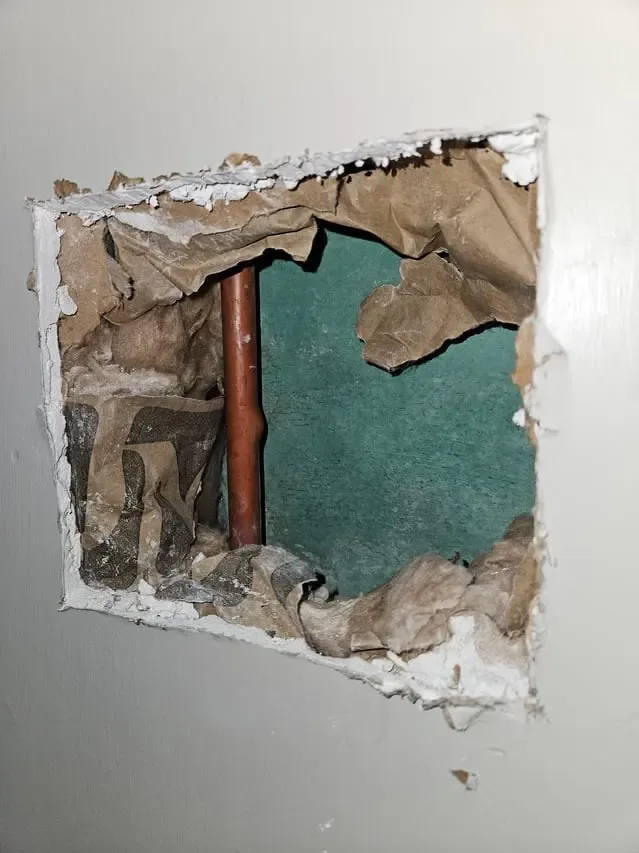 Rectangular drywall opening exposing a copper pipe inside a wall cavity